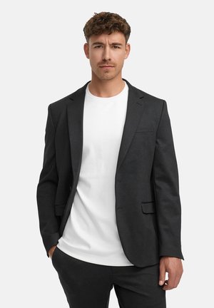 Chaqueta de traje gris oscuro con un ajuste entallado, que presenta solapas de muesca y un solo bolsillo en el pecho, llevada sobre una camiseta blanca lisa.