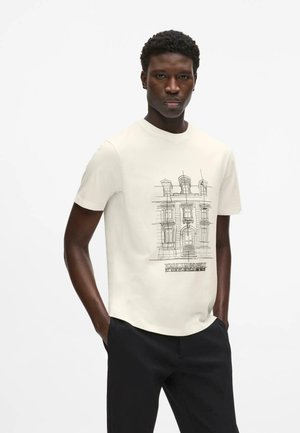 Mann trägt ein weißes T-Shirt mit einer schwarzen Linienzeichnung eines Gebäudes und schwarze Hose, steht mit den Händen in den Taschen vor weißem Hintergrund.