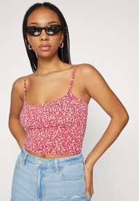 Červený kvetinový crop top s tenkými ramienkami, so zhrnutým dizajnom. Nosený s vysokými modrými denimovými šortkami. Čierne slnečné okuliare pridávajú kontrast.