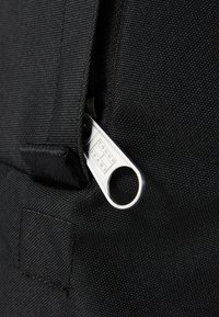 Dettaglio di una borsa di stoffa nera che mette in mostra un tirante in metallo con incisione "TOMMY JEANS", superficie testurizzata e bordo di stoffa cucito a stretto.
