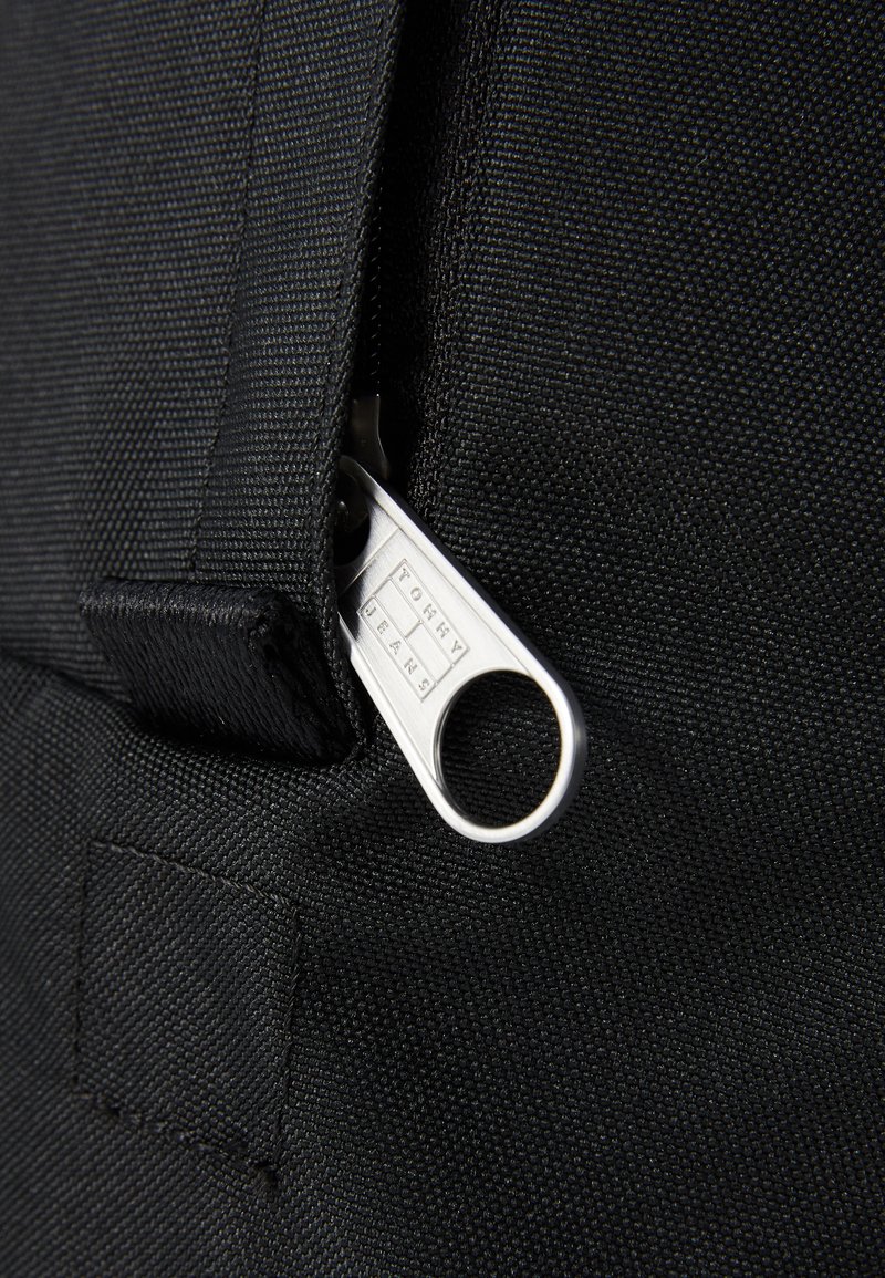 Detail eines schwarzen Stoffbeutels, der einen Metallreißverschluss mit eingravierter "TOMMY JEANS"-Aufschrift, einer strukturierten Oberfläche und einem eng genähten Stoffrand zeigt.