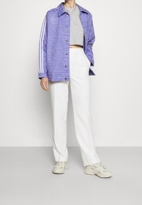 Fialová bunda s bílým vlnkovým vzorem, stříbrné knoflíky, doplněná šedým krátkým topem a bílými kalhotami. Světlé tenisky dokončují vzhled.