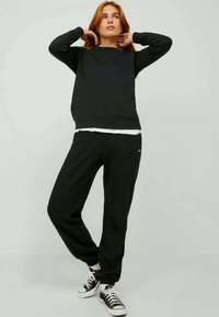 Giovane che indossa un felpa nera, pantaloni della tuta neri e sneakers bianco e nero, in piedi con le mani dietro il collo su uno sfondo semplice.