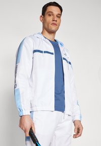 Chaqueta deportiva blanca con acentos azul claro y rayas marino, combinada con una camiseta azul. El modelo sostiene una raqueta de tenis.
