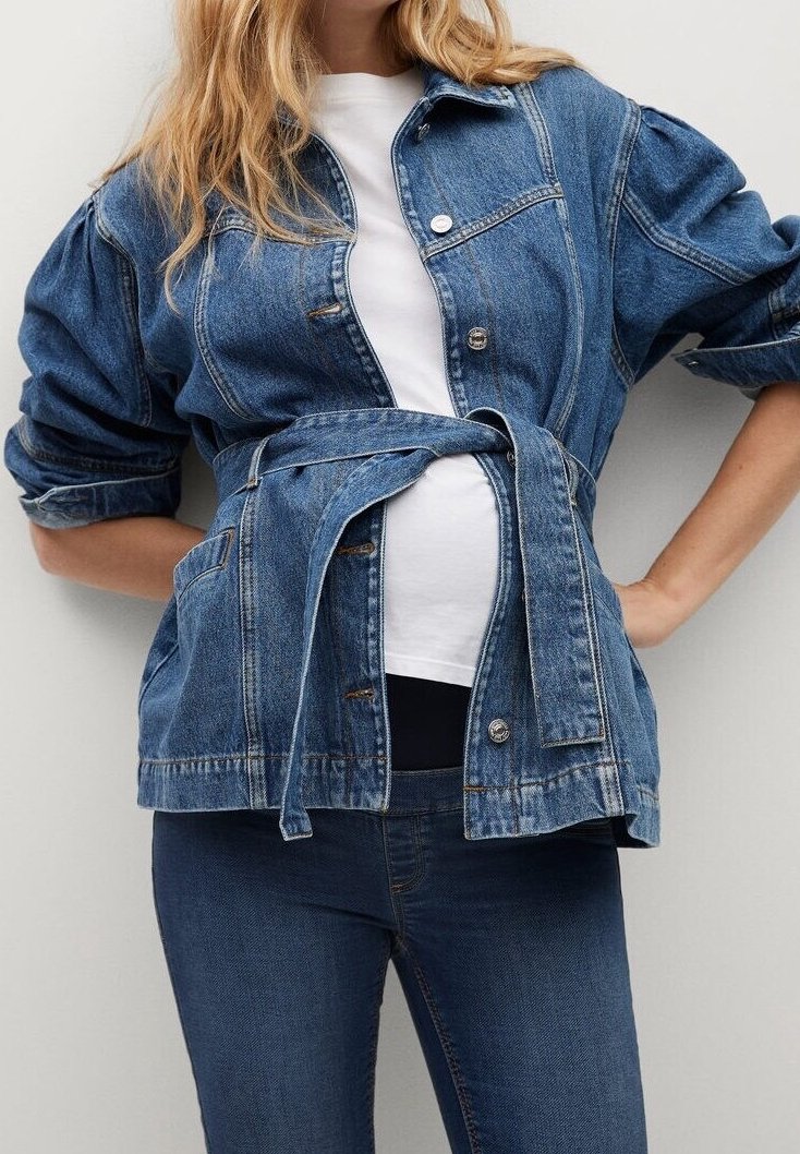 Femme aux longs cheveux blonds portant une veste en jean ceinturée par-dessus une chemise blanche et un jean foncé, debout les mains sur les hanches.