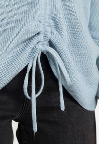 Suéter tricotado azul claro com laterais franzidas, apresentando laços na bainha. Combinado com jeans pretos texturizados, exibindo um corte descontraído.