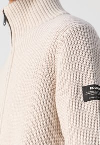 Pull épais tricoté couleur crème avec col montant côtelé et étiquette de marque en tissu noir sur le haut de la manche, avec fermeture éclair.