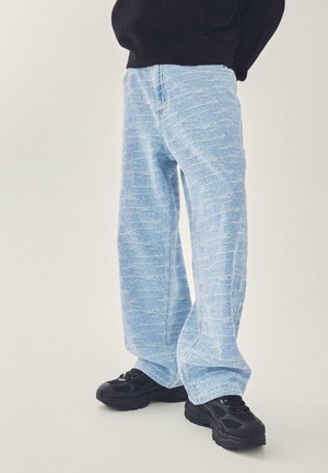 Person trägt hellblaue, strukturierte, weit geschnittene Hosen mit schwarzen klobigen Sneakers und einem schwarzen Oberteil, steht vor einem einfarbigen Hintergrund.