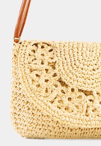 Splotkowana torba na ramię z jasnego żółtego słomy, ozdobiona kwiatowymi wzorami wyciętymi, o teksturowanej powierzchni i brązowym skórzanym pasku.