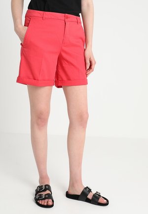Frau trägt leuchtend rote Shorts mit umgeschlagenem Saum, schwarzen Top und schwarze offene Sandalen mit doppelten Schnallenriemen, steht auf weißem Boden.