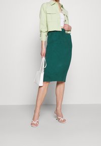 Zelená žebrovaná sukně spárovaná s bledě zelenou zkrácenou bundičkou, bílým topem a bílou kabelkou. Modelka má na sobě bílé sandály na podpatku.