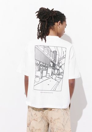 Personne avec des dreadlocks portant un t-shirt blanc avec un dessin en lignes noires représentant une scène de rue en ville et un pantalon beige à motifs.