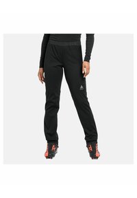 Schwarze Sporthose mit elastischem Bund, glatter Textur und Logodetail auf der linken Seite, kombiniert mit schwarzen und roten Schuhen.
