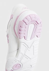 Zapatilla deportiva blanca con un diseño en rosa y gris. La suela cuenta con patrones de agarre texturizados y detalles de marca. Perforaciones transpirables en los laterales.