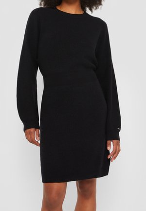 Kobieta ubrana w dopasowaną czarną dzianinową sukienkę z długim rękawem i okrągłym dekoltem, stojąca na tle jednolitego, jasnoszarego tła.