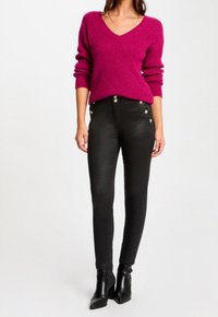 Frau in einem magentafarbenen V-Ausschnitt-Pullover, hochtaillierten schwarzen Hosen mit silbernen Knöpfen und schwarzen, abgesetzten Ankle-Boots vor einem weißen Hintergrund.