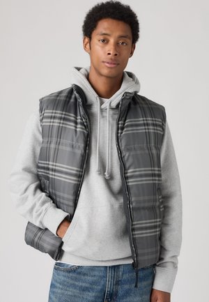 Jeune homme portant une doudoune grise à carreaux par-dessus un sweat à capuche gris clair et un jean bleu, debout avec une main dans la poche devant un fond uni.