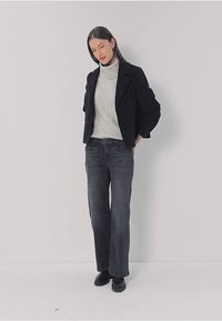 Veste noire courte portée sur un col roulé gris clair, associée à un jean gris foncé taille haute et des bottes noires, sur fond uni.