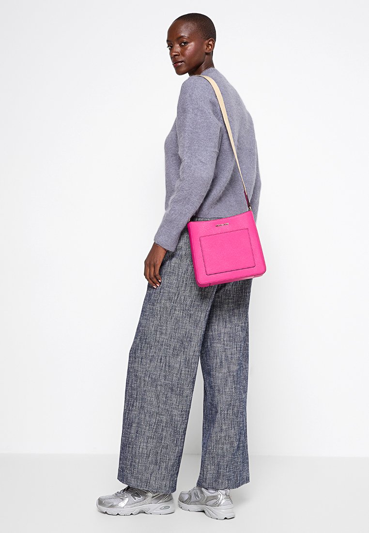 Magenta schoudertas met een gladde textuur, voorzien van een platte voorzak en een afneembare schouderband. G gedragen met een grijze trui en brede broek.