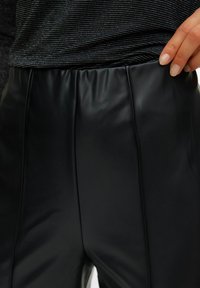 Melnas mākslīgās ādas bikses ar gludu tekstūru, kas aprīkotas ar elastīgu jostu un vertikālām šuvju detaļām priekšpusē.