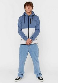 Blauwe en witte capuchontrui met een ritssluiting aan de voorkant, zwarte accenten en zijzakken, gecombineerd met lichtblauwe spijkerbroek en zwarte sneakers.