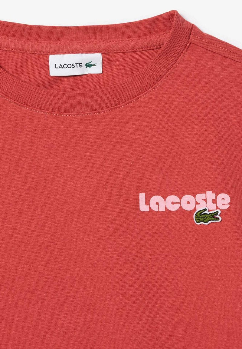Rotes Baumwoll-T-Shirt mit Rundhalsausschnitt, minimalistischer pinker "Lacoste"-Schriftzug und grünem gesticktem Krokodil-Logo auf der linken Seite.