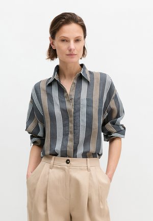 Frau trägt ein dunkel gestreiftes Hemd mit Knöpfen, die Ärmel sind hochgekrempelt, dazu beige, hochgeschnittene Plisseehose mit Taschen, Hände in den Taschen, neutraler Hintergrund.