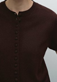 Zbliżenie osoby w ciemnoczerwonym, dzianinowym topie z okrągłym dekoltem i pionowym rzędem guziczków pokrytych materiałem.