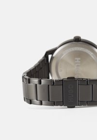 Reloj de pulsera de acero inoxidable negro con una correa lisa, esfera redonda y logo visible en el broche. Presenta un fondo de caja pulido.