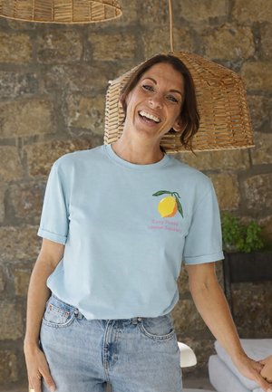 Smiling woman in light blue T-shirt with lemon graphic and "Easy Peasy Lemon Squeezy" text stands indoors with stone wall and wicker lamps.