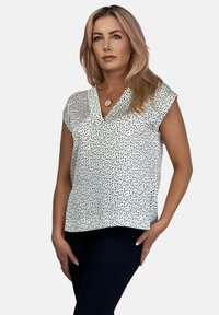 Krótkie rękawy, bluzka z dekoltem w serek w kolorze białym z czarnym, nakrapianym wzorem. Lekka tkanina o luźnym kroju, proste wykończenie dołu i brak dodatkowych ozdób.