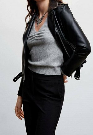 Nainen, joka pukeutunut harmaaseen v-kaulaiseen neulepaitaan, mustaan nahkatakkiin, mustiin housuihin ja hopeiseen ketjukaulakoruun, yksinkertaisen taustan edessä.