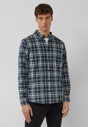 Młody mężczyzna w granatowo-białej kraciastej koszuli zapinanej na guziki i czarnych spodniach, stojący na tle jednolitego jasnoszarego tła.