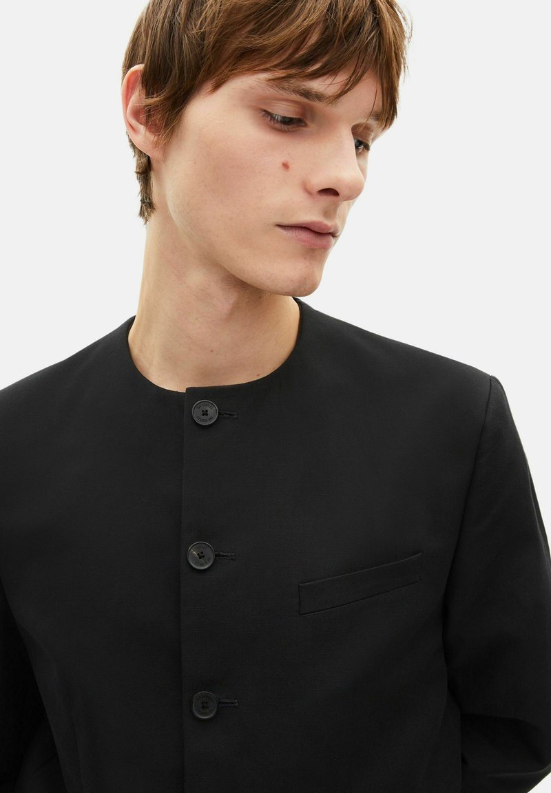 Black buttoned jacket featuring a round neck, structured fit, and a left chest pocket. Fabric appears smooth and slightly textured.