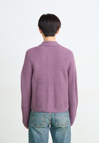 Lila gestrickter Pullover mit gerippter Textur und kurzem Kragen, kombiniert mit hellblauen Jeans. Die Rückenansicht betont die Passform des Pullovers.