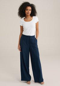 Weißes Kurzarmoberteil kombiniert mit marineblauen weiten Hosen. Die Hose verfügt über einen Kordelzugbund und eine gefaltete Front. Das Model trägt Sandalen.