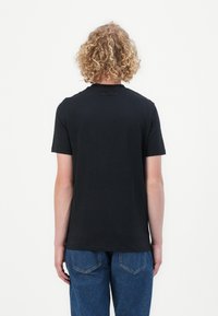 Fekete, rövid ujjú póló, kerek nyakkivágással, puha pamutból készült. Diszkrét logó található a nyaknál. Kék farmernadrággal viselt.