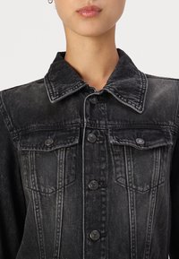 Črno denim jakna z klasifikatorjem ovratnikom, gumbom na sprednji strani, dvema žepoma na prsih in poškodbami teksture z vidnimi šivi.
