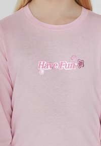 Różowa bluzka z długim rękawem z napisem "Miej Zabawę" w błyszczących, metalicznych literach z akcentami w kształcie gwiazd. Miękka tekstura i klasyczny okrągły dekolt.