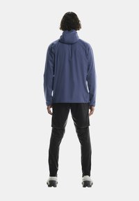 Giacca impermeabile blu con cappuccio, abbinata a pantaloni cargo neri. Include un taglio comodo e elementi regolabili per il massimo comfort.
