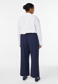 Žena s tmavými vlasy svázanými do drdolu, oblečená v bílé košili s dlouhým rukávem, tmavě modrých širokých kalhotách a tmavě modrých balerínách, stojící zády k nám.