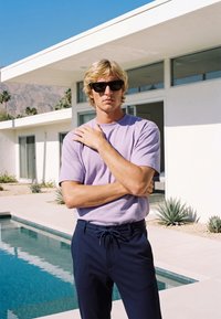 Jeune homme aux cheveux blonds et lunettes de soleil, bras croisés, debout près d'une piscine moderne et d'une maison blanche sous un ciel bleu clair.