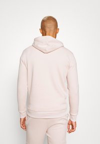 Felpa rosa chiaro con tasca a marsupio, texture morbida e cappuccio con cordino regolabile; indossata sopra pantaloni della tuta abbinati.