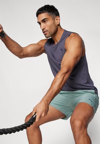 Haut de sport sans manches violet, shorts vert clair, silhouette musclée, tenant une corde d'entraînement noire, fond uni.