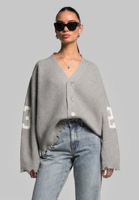 Grey, ribbed cardigan with a V-neck, three buttons, and patch detailing. Features frayed hems and long sleeves, paired with light blue jeans.