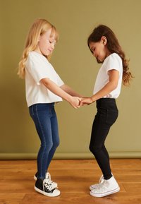 To børn holder hinandens hænder og bærer hvide t-shirts. Den ene har blå jeans og sorte sneakers, mens den anden har sorte jeans og hvide sneakers.