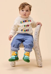Bambino che indossa un maglione lavorato a maglia color crema con testo colorato, jeans blu e scarpe multi-strappo verdi e gialle, seduto su una sedia maculata.