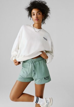 Kvinde med krøllet hår iført en hvid Red Bull Racing sweatshirt og grønne Red Bull Racing shorts, stående på ét ben med hvide sneakers.