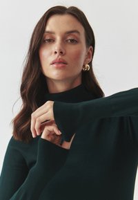 Tamnozelena turtleneck dukserica s rebrastim rukavima. Model ističe teksturu tkanine, prikazujući prilagođeni dizajn i duge rukave.