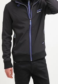 Svart softshell-jacka med blå dragkedjeaxlar, huva och fickor. Den har en figursydd design med ribbad krage och ärmar.